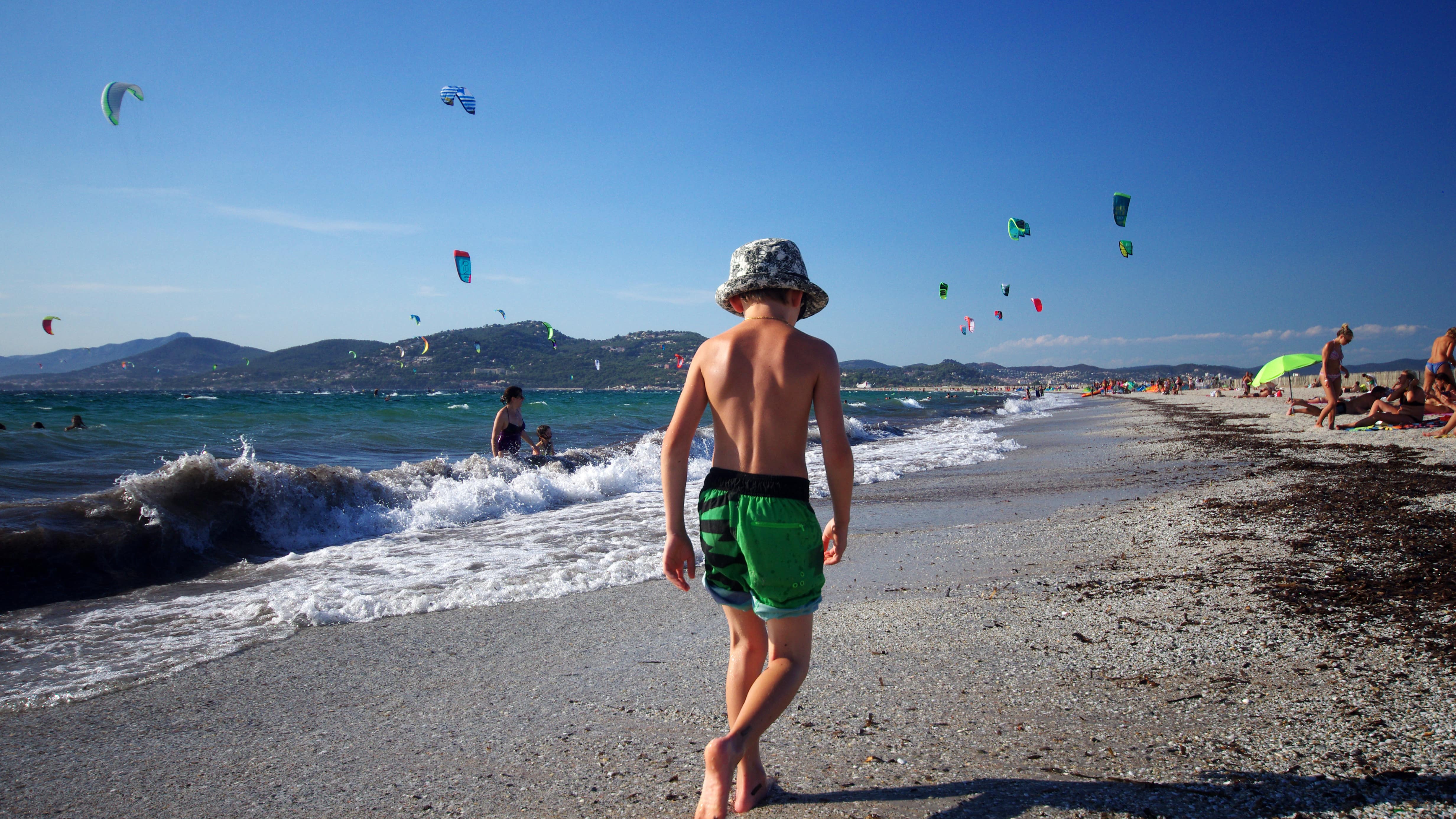 Enfant de dos, au bord de l’eau, des familles installées sur le sable et des voiles de kitesurf sur la plage de l’Almanarre