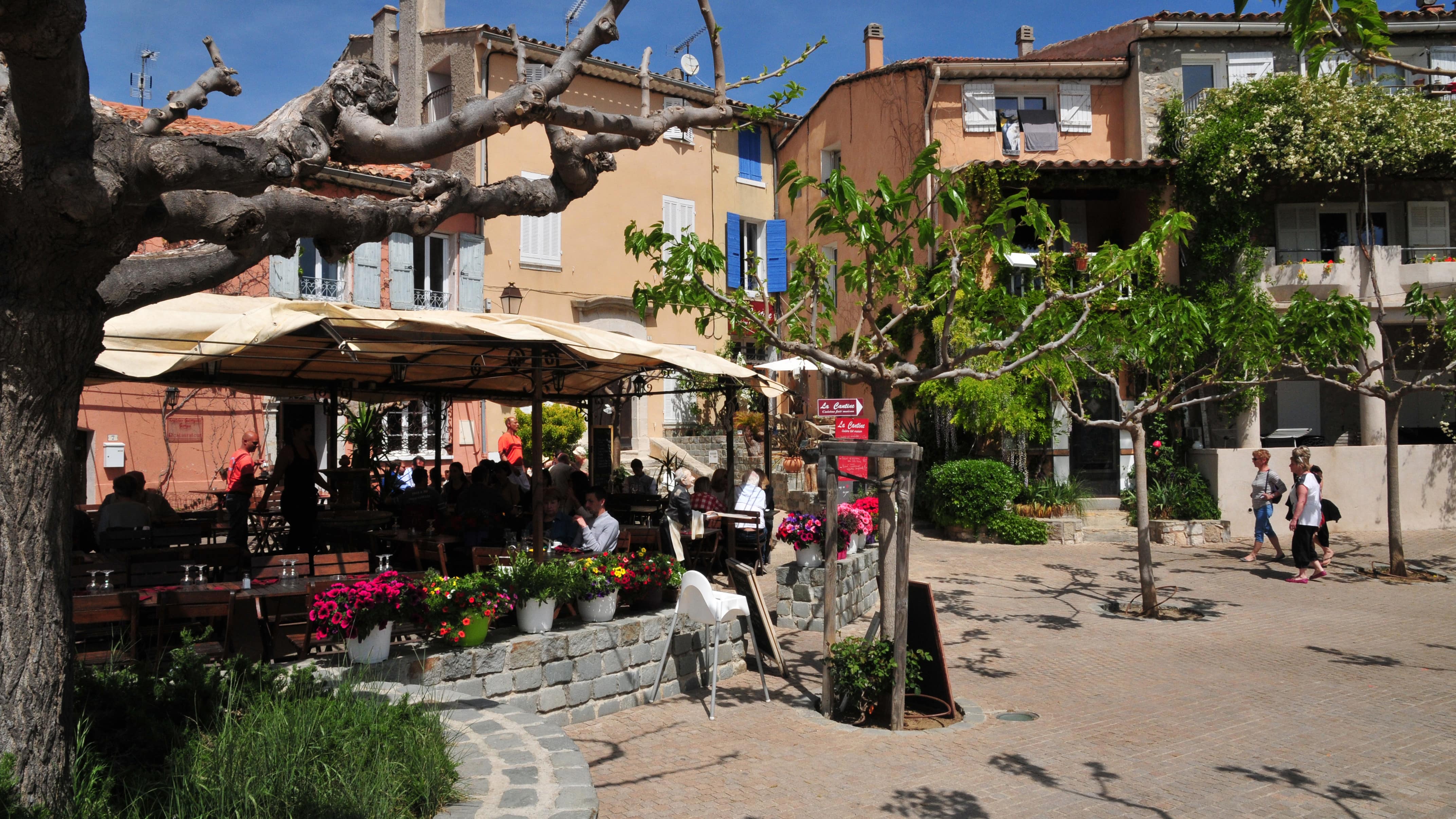 Quelques promeneurs sur une place et des personnes attablées sur la terrasse ombragée d’un restaurant au Castellet dans le Var